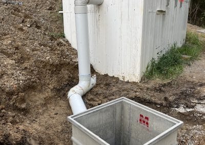 Construction site with a large concrete foundation wall, white PVC pipes, and a dirt trench; a gray plastic bin sits in the foreground.