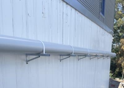 Long white utility pipe runs horizontally along a white wall, supported by metal brackets beneath a corrugated upper facade.