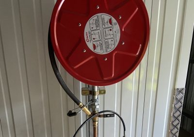 Red wall-mounted hose reel with black hose and copper piping, valves, and a Ridgid toolbox on a diamond-plate wall in a workshop setting.