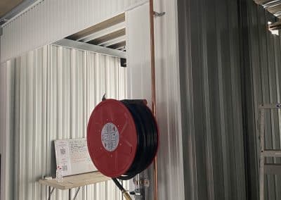 Copper pipe running vertically along a corrugated metal wall in a workshop, with a red hose reel mounted nearby and a small work desk in the background.