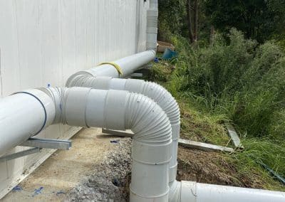 Grey PVC downspouts and large curved drainage pipes along the side of a white building, with dirt and plants nearby.