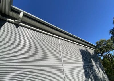 Side of a corrugated metal building with white gutters and downspouts along the roof, set against a clear blue sky and tree shadows.