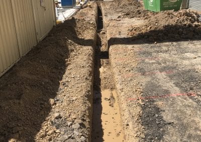 Deep construction trench with muddy water and piled dirt beside an industrial building, with equipment in the background and a portable toilet present.