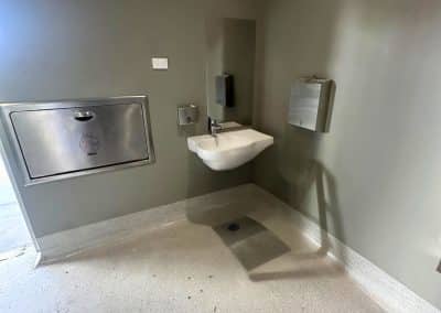 Public restroom corner with a small wall-mounted sink, mirror, soap dispenser, and metal hand-dryer/hand-dryer unit on gray walls; ventilation grille above.
