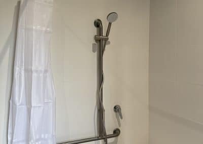Shower area with white tiled walls, a white curtain on a rod, and a chrome handheld shower near a wall-mounted faucet above a bench-like seat.