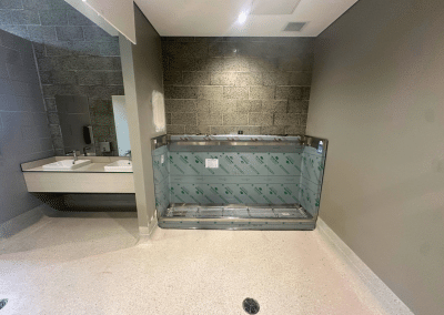 Public restroom area under construction with two sinks on the left, gray tile walls, and a stainless steel fixture wrapped in protective film at center; floor drain visible in foreground.