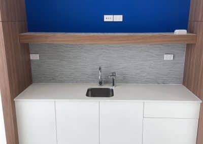 Modern kitchen with a blue accent wall, wooden trim, grey tile backsplash, and white cabinets beneath a stainless steel sink.