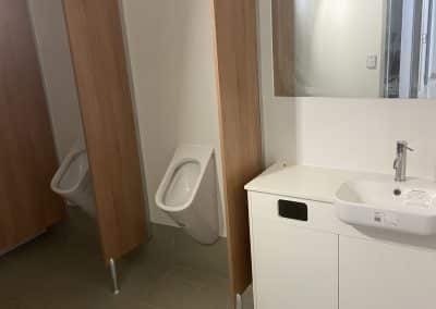 Public restroom with wooden stall dividers, urinals, a white sink vanity, and a large mirror on white tiled walls.