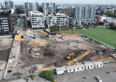Construction site with excavators and dirt piles, surrounded by modern apartment buildings and a green soccer field in the background.