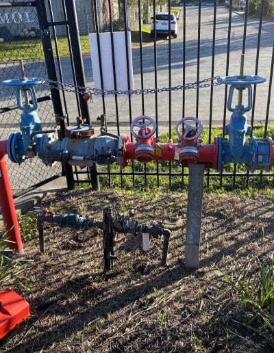 Industrial pipe manifold with blue gate valves and red main pipe behind a chain-link fence.