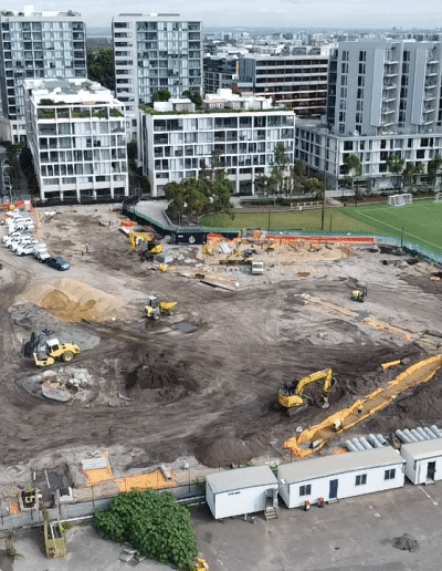 Construction site with excavators and dirt piles, surrounded by modern apartment buildings and a green soccer field in the background.