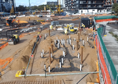 Active construction site with numerous vertical pipes and worker crews preparing foundation in a fenced area near a roadway and modern buildings in the background