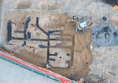 Aerial view of an active construction site with a network of excavated trenches and piles of dirt, plus a small yellow excavator and scattered pipes.