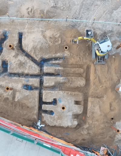 Aerial view of an active construction site with a network of excavated trenches and piles of dirt, plus a small yellow excavator and scattered pipes.