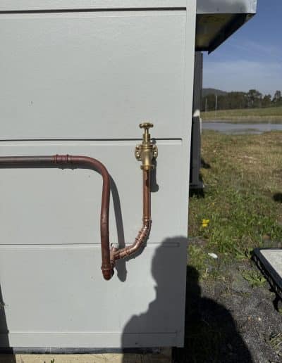 Outdoor utility cabinet with copper water line and brass shutoff valve outside a grey metal enclosure.