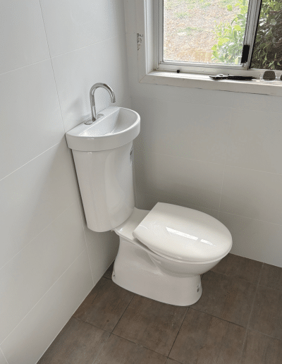 Small bathroom with a white toilet and a compact corner sink featuring a curved chrome faucet; white tiled walls and a window above showing greenery outside. On the window sill are a few items (sunglasses,工具等).