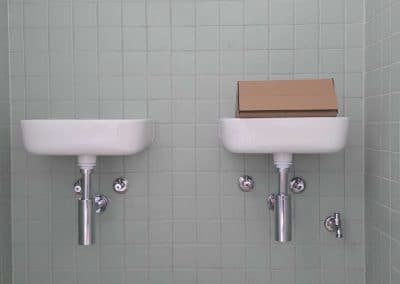 Two white wall-mounted sinks with chrome pipes on a pale green tiled wall; a brown box sits on the right sink.