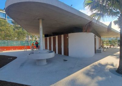 Modern circular concrete canopy over a building entrance, supported by a column, with a palm tree on the right and construction workers in the background.