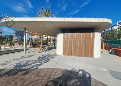 Open-air construction site with a large curved concrete canopy, wooden slat wall, and clear blue sky behind.