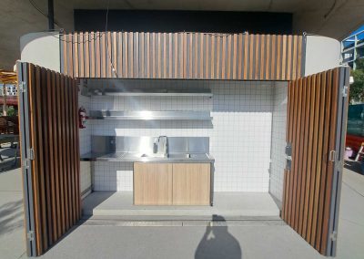 Outdoor kitchen station enclosed by vertical wooden slats, featuring a stainless steel sink and counter against a white tiled wall, under a concrete overhang.