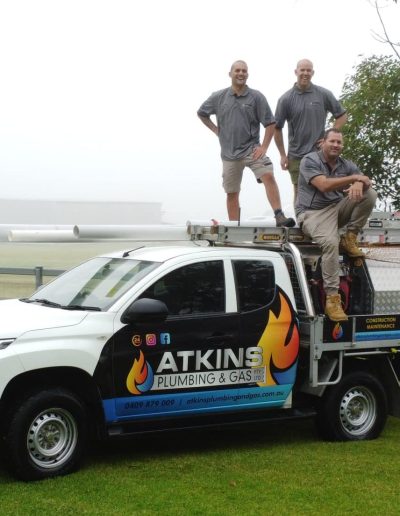 Five workers in gray uniforms pose on and around a white service truck labeled Atkins Plumbing & Gas in a foggy, grassy field.