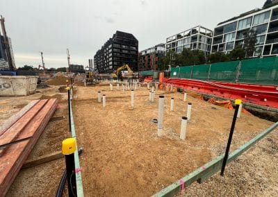 Construction site with PVC pipes, dirt, and machinery, set against modern apartment buildings in the background.