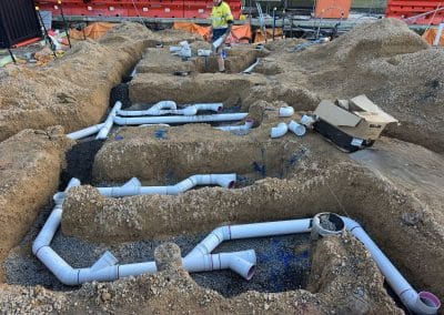 Construction site with multiple trenches and white PVC drainage pipes; a worker in a yellow safety vest inspects the area.