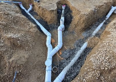 Construction workers laying large white PVC pipes in a deep trench at a construction site, with dirt piles, tools, and safety gear nearby.