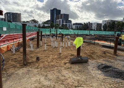 Construction site with orange barriers, green safety fencing, and white PVC pipes arranged in the dirt foreground, urban buildings in background.