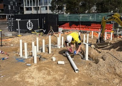Construction worker in a yellow safety vest and white helmet bending over PVC pipes at a dirt construction site, with modern apartment buildings in the background