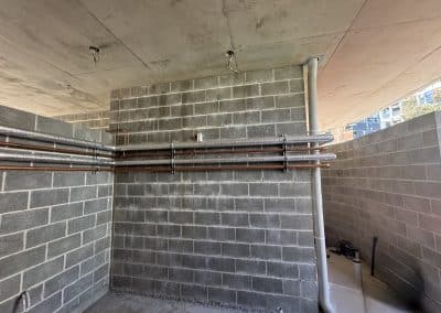 Unfinished utility space with gray concrete block walls, exposed pipes along the wall, and hanging electrical wires from the ceiling.
