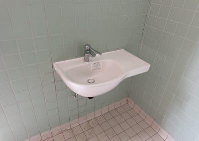 White wall-mounted sink with a chrome faucet against pale green tiled walls; exposed pipes beneath.