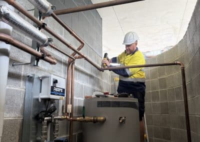 Worker in yellow safety shirt and white hard hat uses a drill beside a tall water heater and copper piping in a utility room.