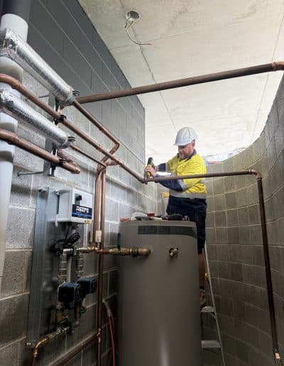 Worker in yellow safety shirt and white hard hat uses a drill beside a tall water heater and copper piping in a utility room.