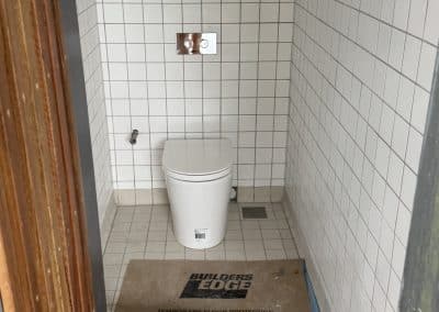 Small, unfinished bathroom with white square tiles, a toilet, and a wall flush plate; a Builders Edge temporary floor protector lies on the floor.
