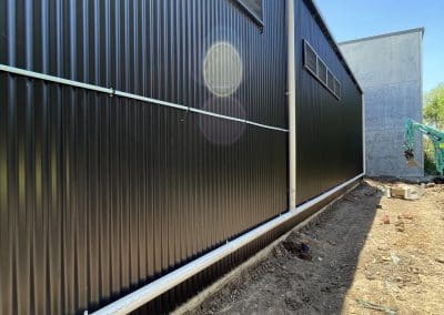 Side view of a black corrugated metal building with white utility pipes running along the foundation under a clear blue sky, construction site nearby.