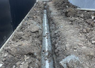 Trench dug between a dark vertical wall and a light blue wall, with a concrete pipe line in the center and piled dirt on both sides for drainage work