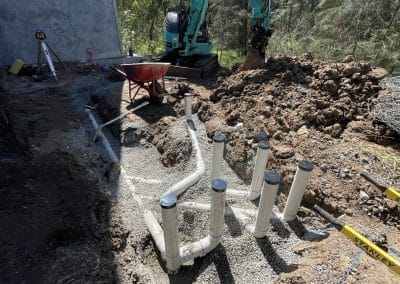 Teal compact excavator digging a trench beside a building, with white PVC pipes and a dirt mound nearby.