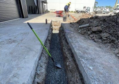 Construction worker in a high-visibility shirt shovels gravel into a deep narrow trench beside a building under construction.