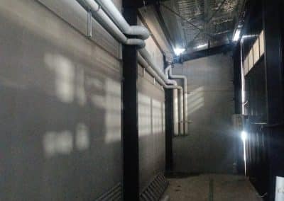 Industrial service corridor with white pipes running along wall and ceiling, supported by black metal beams, dim lighting nearby.