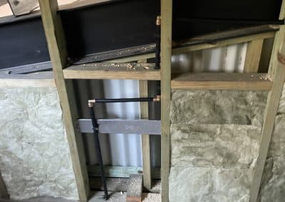 Construction space with wooden studs, metal framing, and exposed pipes behind a black beam or panel; dust and debris on the concrete floor.