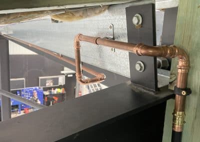 Copper plumbing pipes and elbows attached to a metal beam under a wooden structure in a workshop/industrial setting.