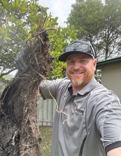 Smiling man in a gray work shirt and cap stands next to a large uprooted tree with exposed roots in a backyard.