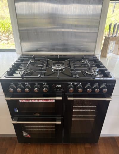 Black stainless steel gas range with multiple burners, metal vent hood behind, and a knife block to the right on a light countertop; sign reads 'DO NOT LIFT BY HANDLES' on the oven door.