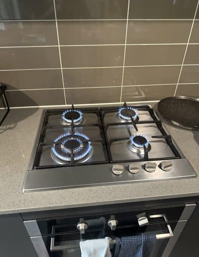 Stainless steel gas stove with four burners burning blue flames, on a speckled countertop with a tiled backsplash behind.