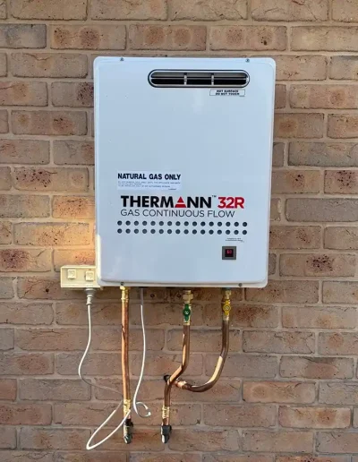 Exterior natural gas water heater (Thermann) mounted on a brick wall with copper gas pipes and a power cord visible.