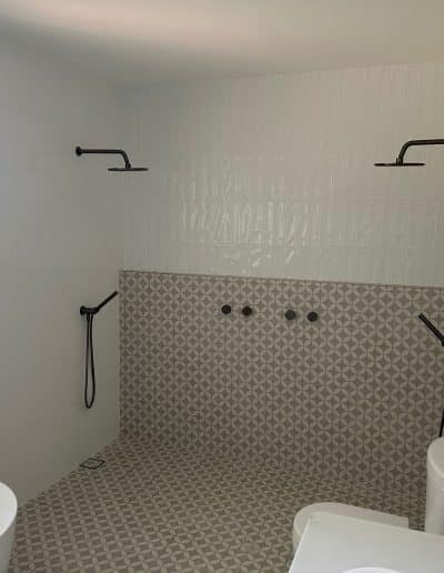 Bathroom with two wall-mounted rainfall showerheads, grey geometric floor tiles, white walls, and a window to the left.
