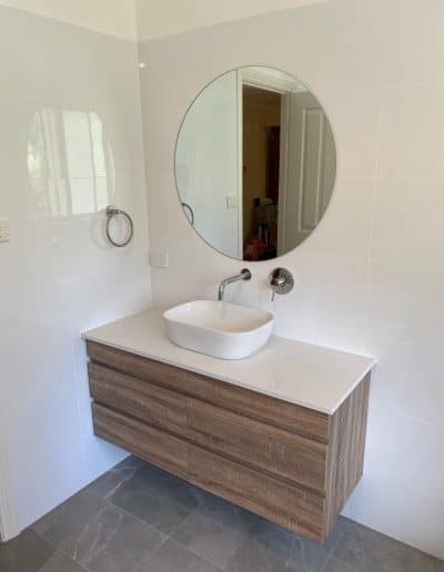 Round mirror above a white ceramic vessel sink on a wood-toned vanity in a bright, minimalist bathroom.
